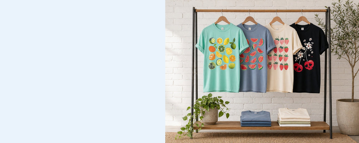4 printed garment-dyed cotton t-shirts on wooden hangers on a wood and iron clothing rack against a sunlit white brick wall, with an olive tree, trailing pothos and folded tees on the shelf below
