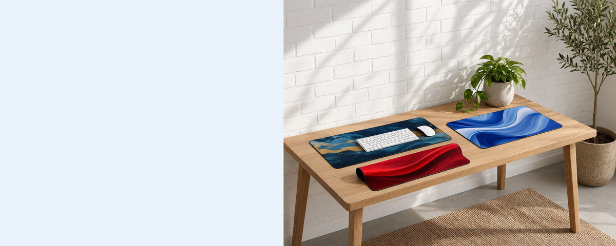 Three printed neoprene desk mats on a tidy desk against a sunlit white brick wall, one in use under a keyboard, one lying flat beside it and one loosely rolled on the desk, with an olive tree