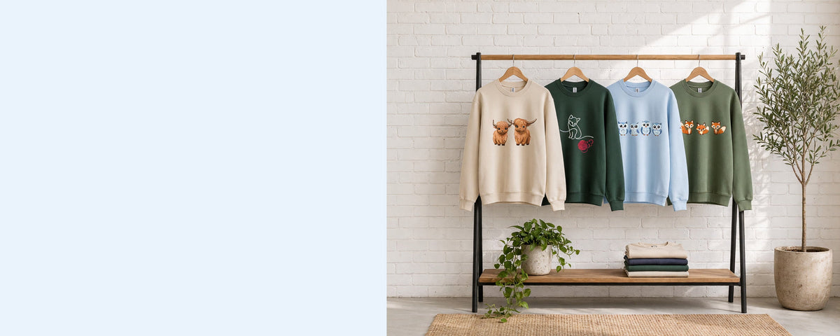 4 printed crewneck sweatshirts on wooden hangers on a wood and iron clothing rack against a sunlit white brick wall, with an olive tree, trailing pothos and folded tees on the shelf below
