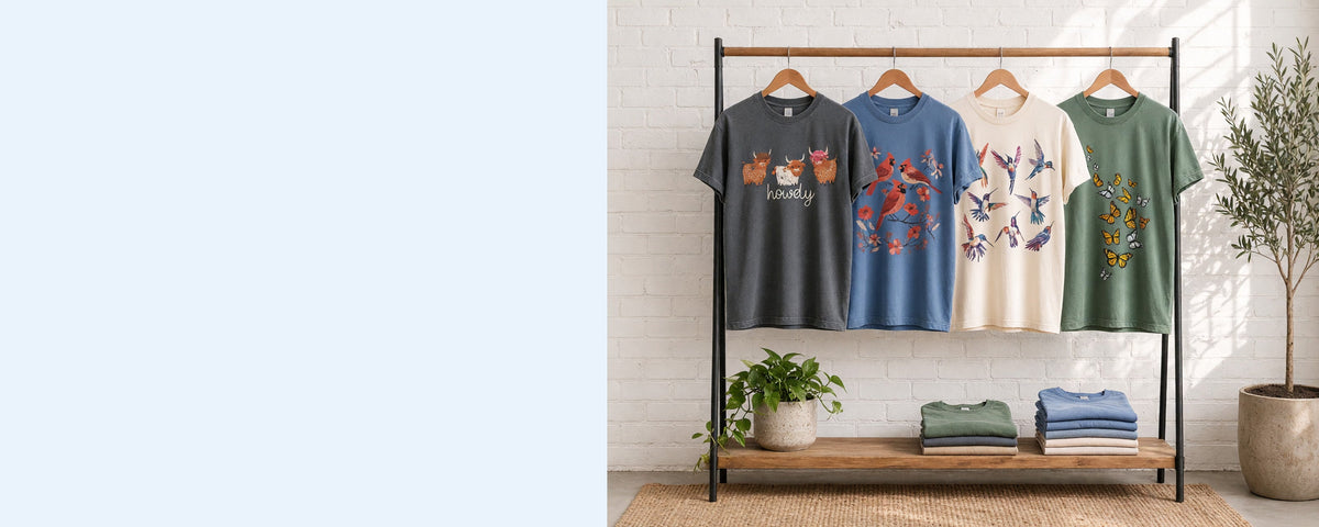 4 printed garment-dyed cotton t-shirts on wooden hangers on a wood and iron clothing rack against a sunlit white brick wall, with an olive tree, trailing pothos and folded tees on the shelf below