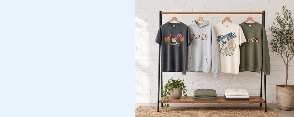 4 printed garments on wooden hangers on a wood and iron clothing rack against a sunlit white brick wall, with an olive tree, trailing pothos and folded tees on the shelf below