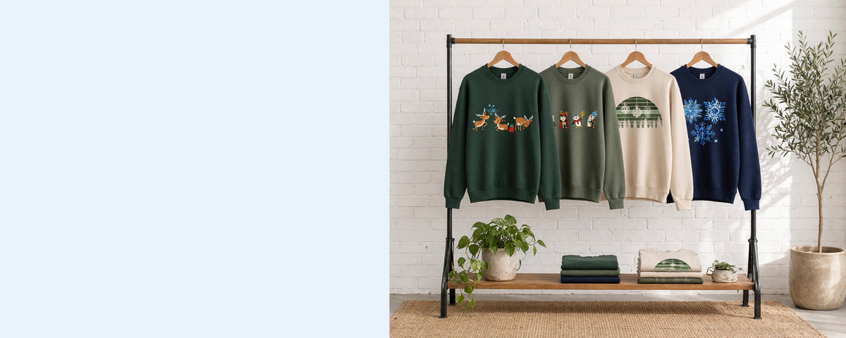4 printed crewneck sweatshirts on wooden hangers on a wood and iron clothing rack against a sunlit white brick wall, with an olive tree, trailing pothos and folded tees on the shelf below