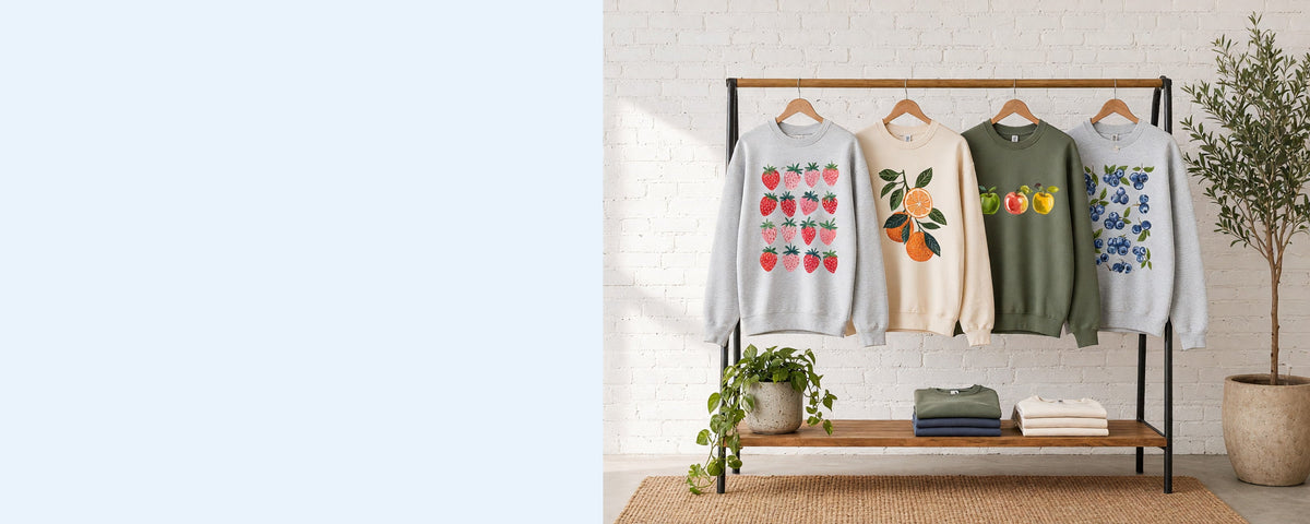 4 printed crewneck sweatshirts on wooden hangers on a wood and iron clothing rack against a sunlit white brick wall, with an olive tree, trailing pothos and folded tees on the shelf below