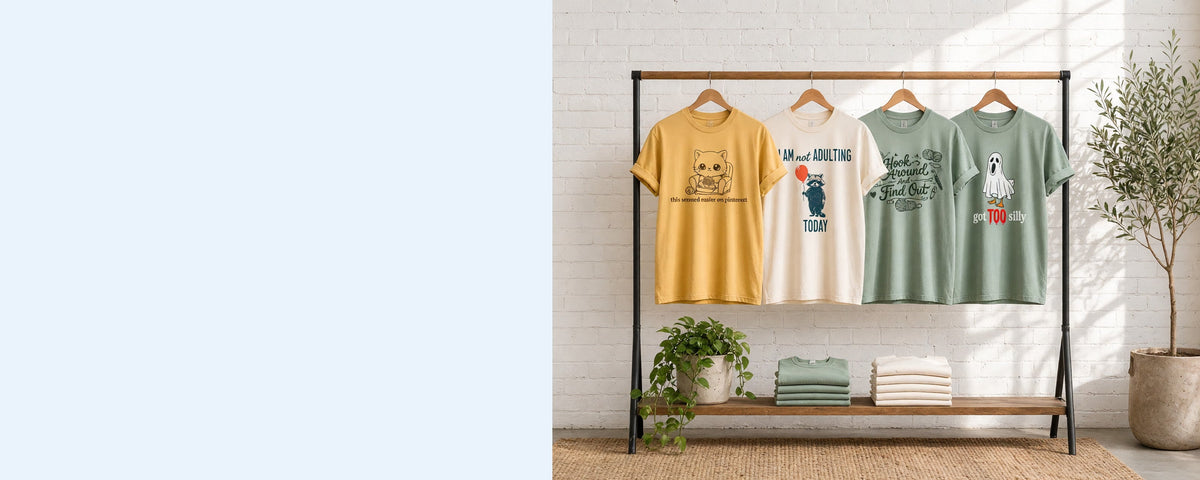 4 printed garment-dyed cotton t-shirts on wooden hangers on a wood and iron clothing rack against a sunlit white brick wall, with an olive tree, trailing pothos and folded tees on the shelf below