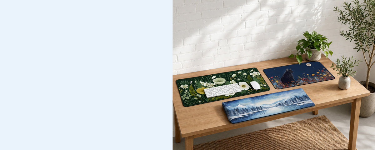 Three printed neoprene desk mats on a tidy desk against a sunlit white brick wall, one in use under a keyboard, one lying flat beside it and one loosely rolled on the desk, with an olive tree