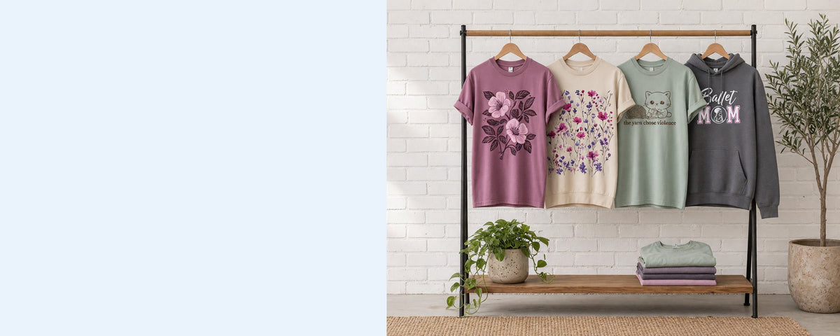4 printed garments on wooden hangers on a wood and iron clothing rack against a sunlit white brick wall, with an olive tree, trailing pothos and folded tees on the shelf below