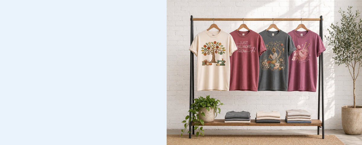 4 printed garment-dyed cotton t-shirts on wooden hangers on a wood and iron clothing rack against a sunlit white brick wall, with an olive tree, trailing pothos and folded tees on the shelf below