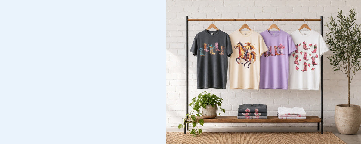 4 printed garment-dyed cotton t-shirts on wooden hangers on a wood and iron clothing rack against a sunlit white brick wall, with an olive tree, trailing pothos and folded tees on the shelf below