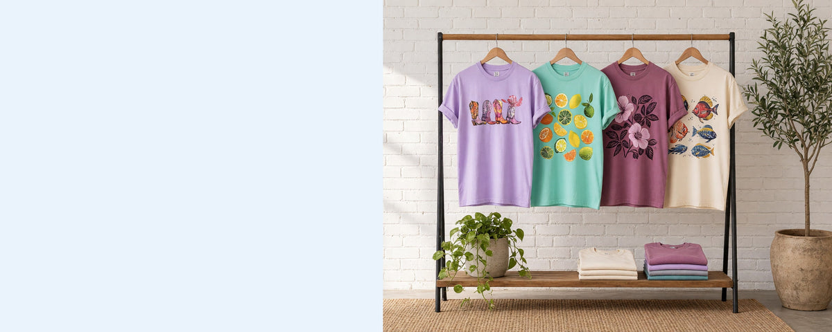4 printed garment-dyed cotton t-shirts on wooden hangers on a wood and iron clothing rack against a sunlit white brick wall, with an olive tree, trailing pothos and folded tees on the shelf below