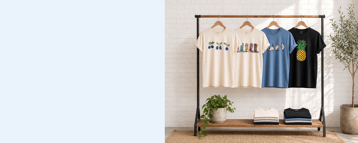 4 printed lightweight cotton t-shirts on wooden hangers on a wood and iron clothing rack against a sunlit white brick wall, with an olive tree, trailing pothos and folded tees on the shelf below