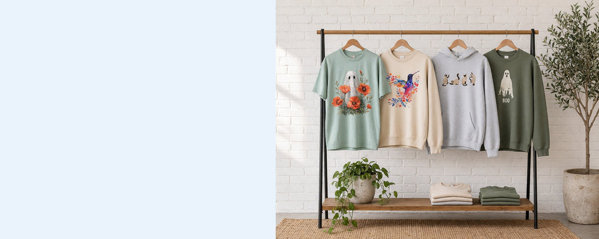 4 printed garments on wooden hangers on a wood and iron clothing rack against a sunlit white brick wall, with an olive tree, trailing pothos and folded tees on the shelf below