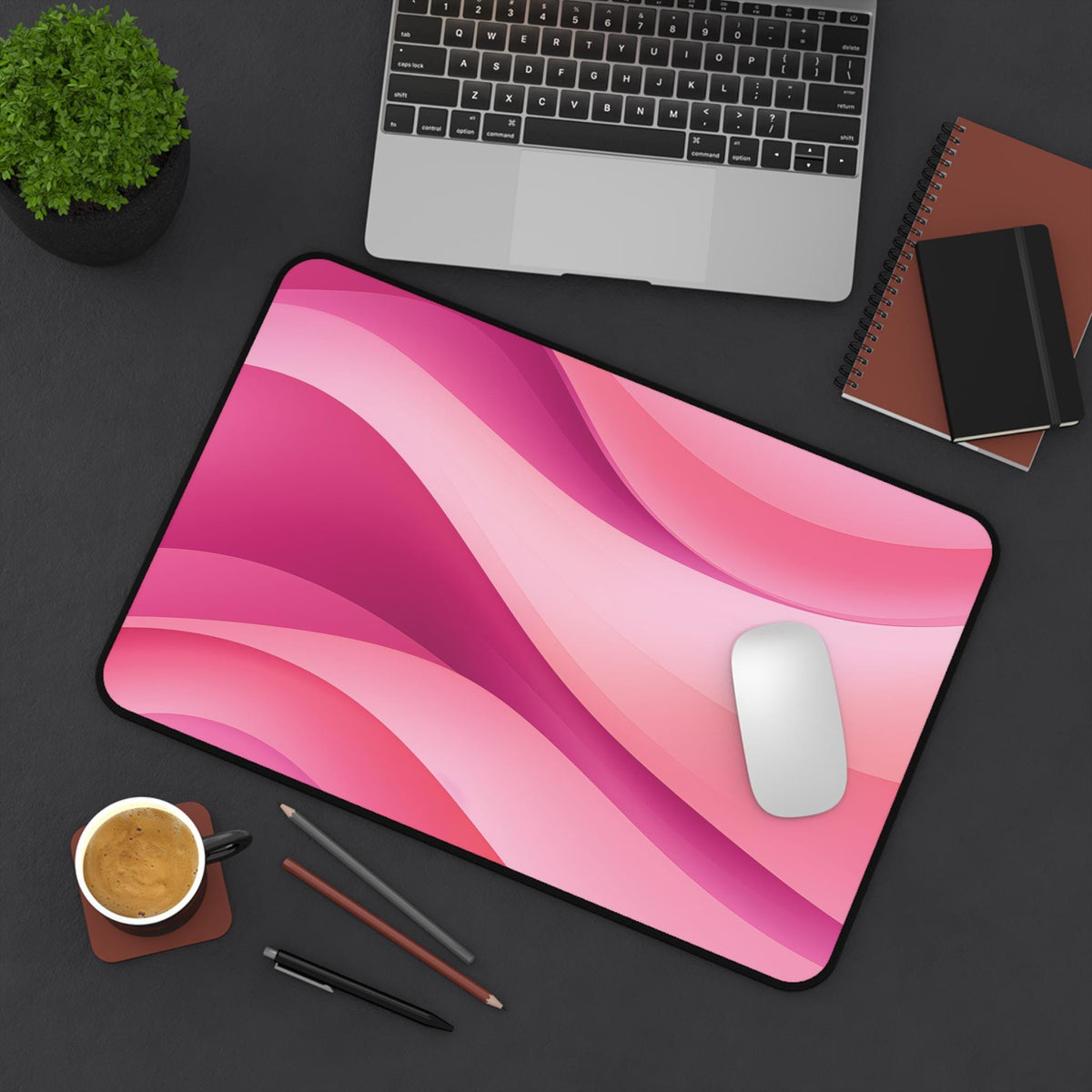 Abstract Desk Mat in pink on dark desk with laptop, plant, notebooks, coffee, pens, mouse from above, 12 × 18 inches.