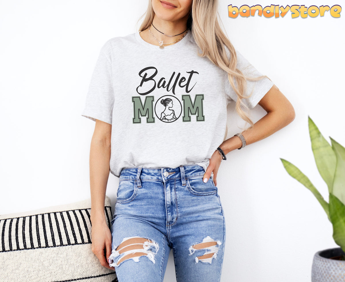Ballet Mom T-Shirt in ash on a woman leaning by a white wall beside a striped pillow and plant.