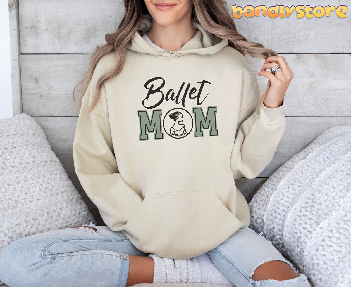 Ballet Mom Hoodie in sand on woman sitting cross-legged amid white pillows by wood panel wall.