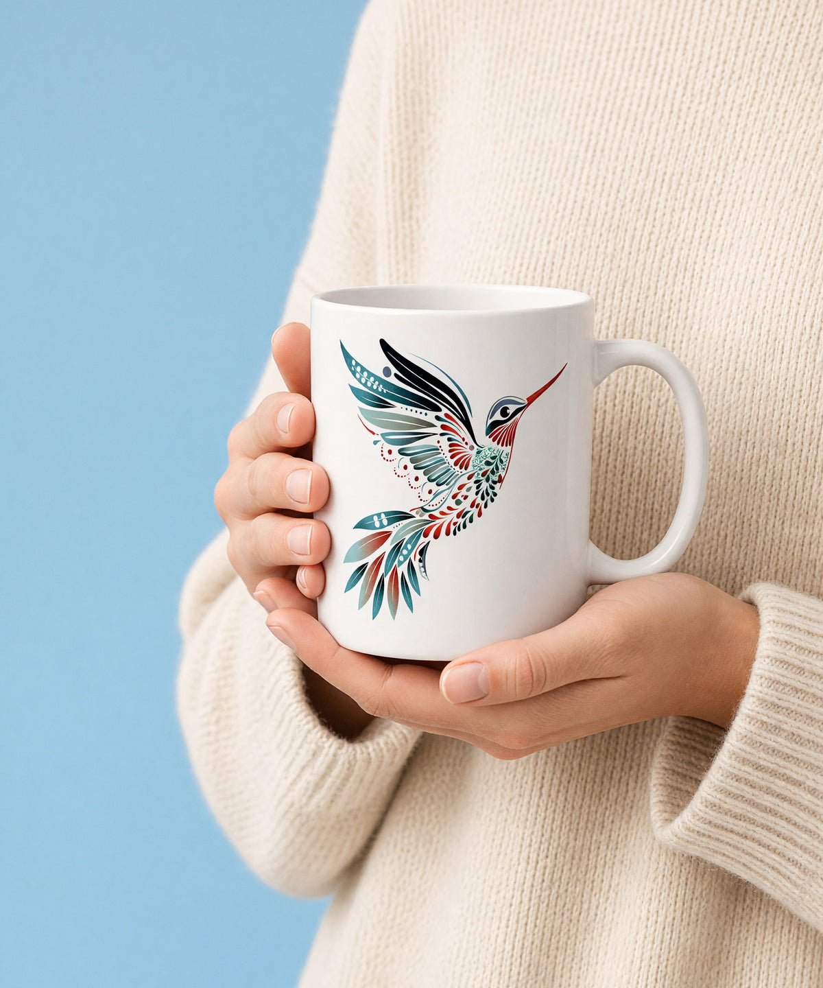 Two hands in a cream knit sweater cradling a white ceramic mug printed with a folk-art hummingbird, against a sky-blue backdrop