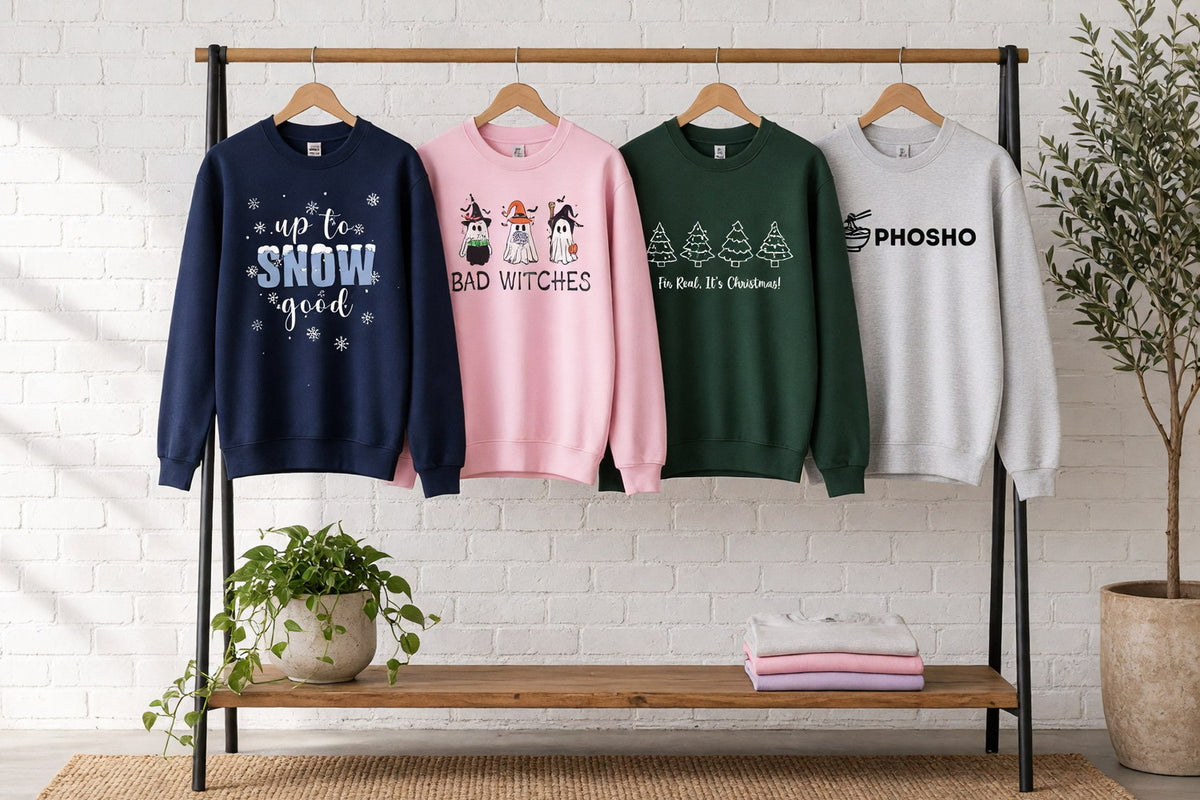 Four funny crewneck sweatshirts with Up To Snow Good, Bad Witches ghosts, Fir Real It's Christmas and Pho Sho designs on wooden hangers against a sunlit white brick wall