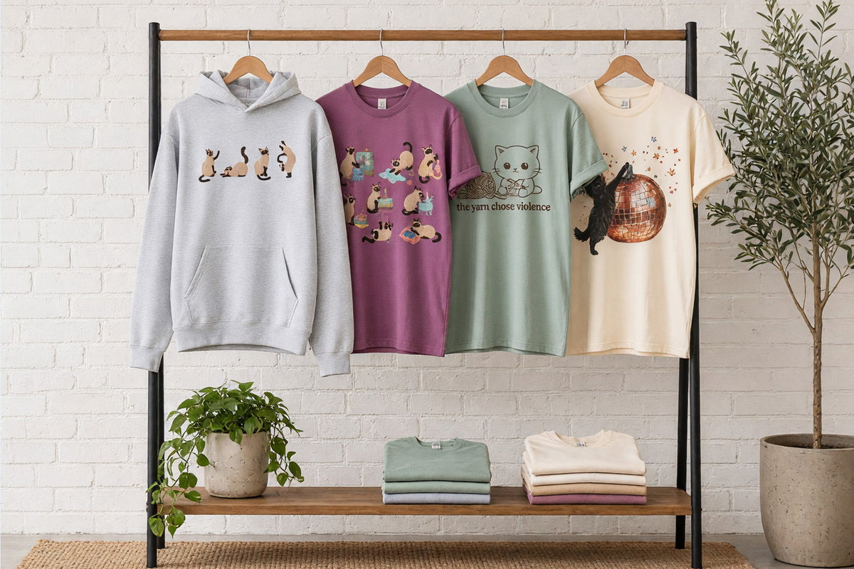 Cat-design tees and sweatshirts on wooden hangers on a clothing rack against a sunlit white brick wall