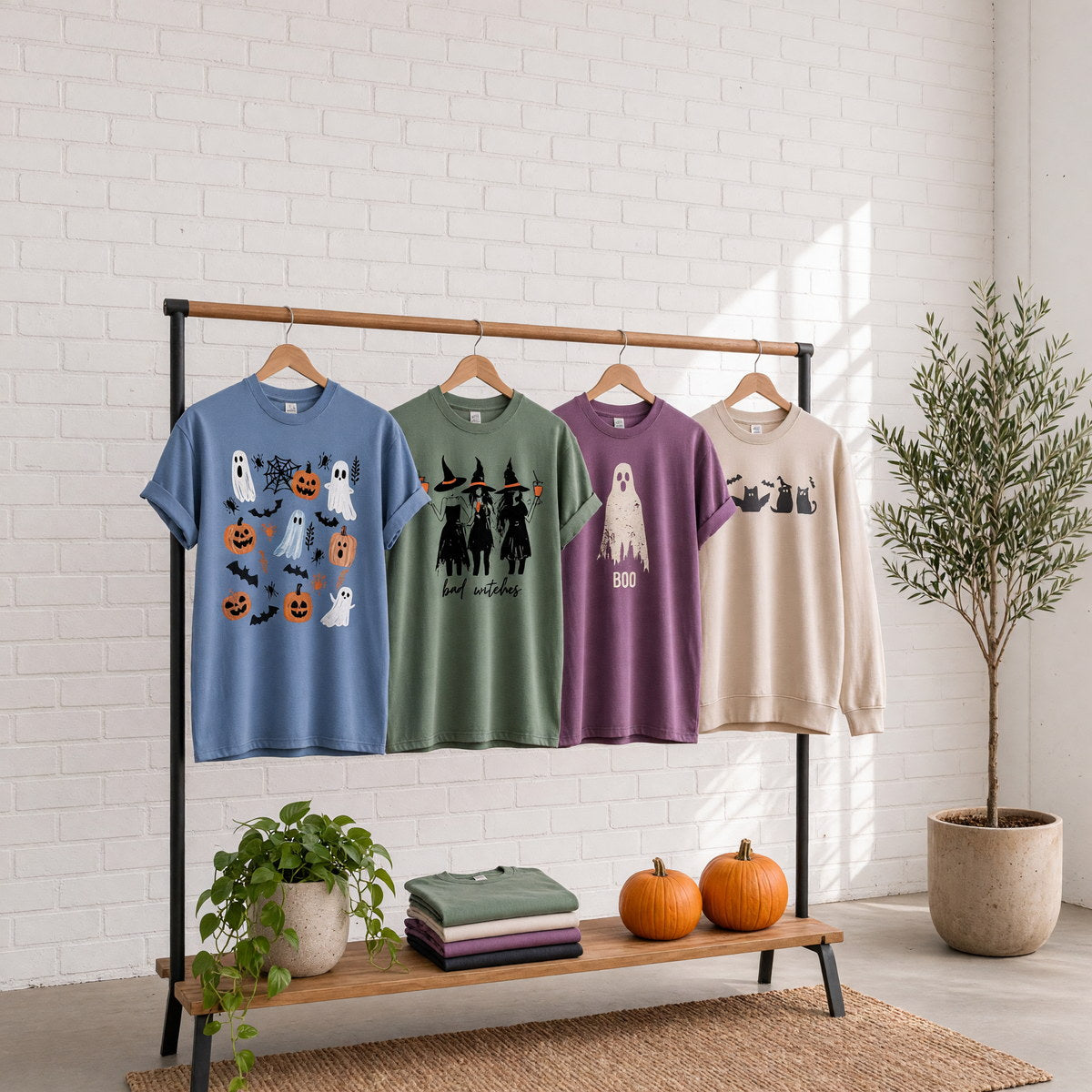 Four printed Halloween t-shirts and sweatshirts on wooden hangers on a wood and iron clothing rack against a sunlit white brick wall with pumpkins on the shelf, empty wall above the rack