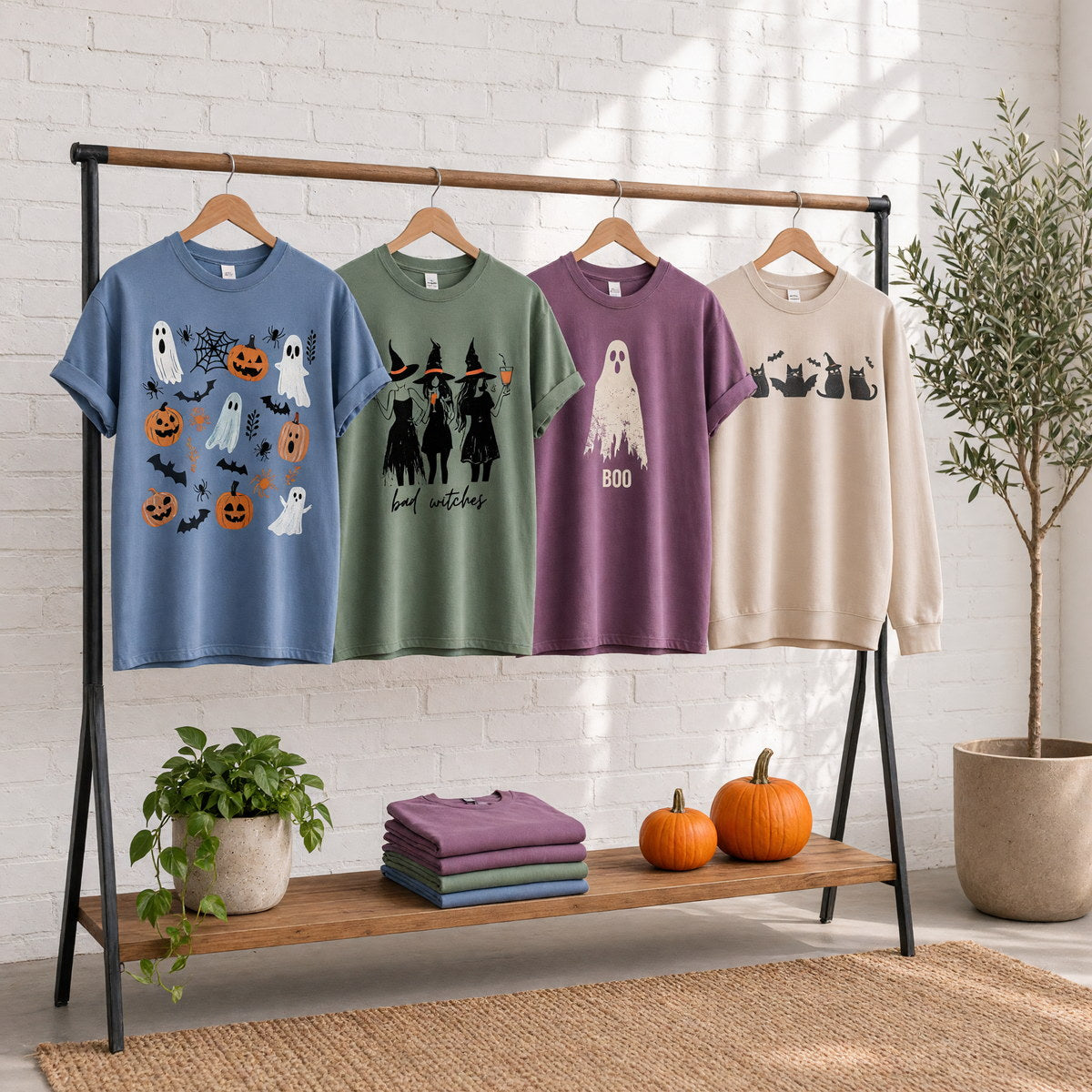 Four Halloween printed garments on wooden hangers on a wood and iron clothing rack against a sunlit white brick wall, with an olive tree, trailing pothos, folded tees and two small pumpkins on the shelf below