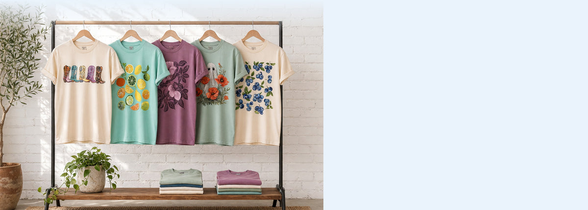 Five printed cotton t-shirts on wooden hangers on a wood and iron clothing rack against a sunlit white brick wall, with an olive tree, trailing pothos and folded tees on the shelf below
