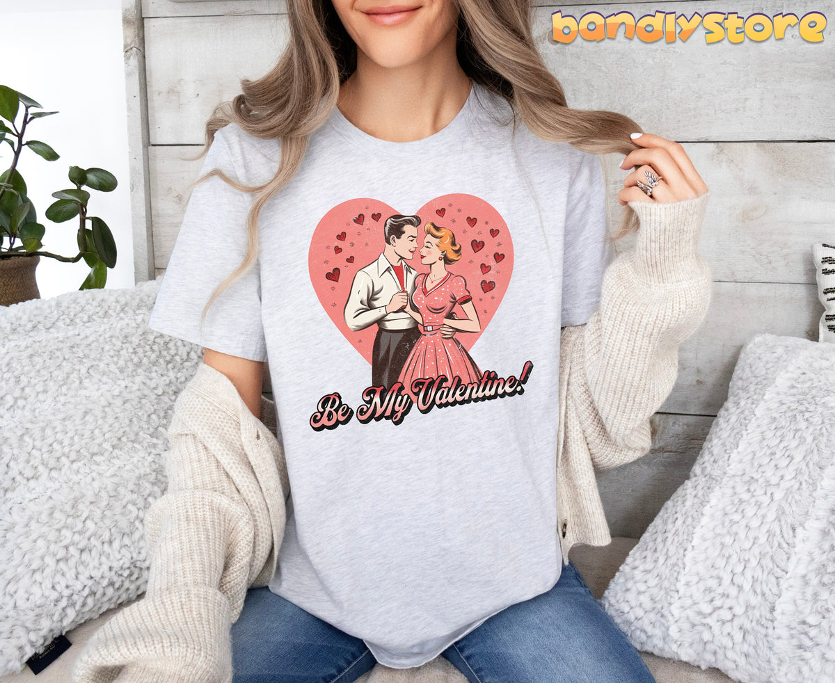 Be Mine Valentine T-Shirt in ash on a woman seated front-facing amid white pillows, plant and wooden wall.