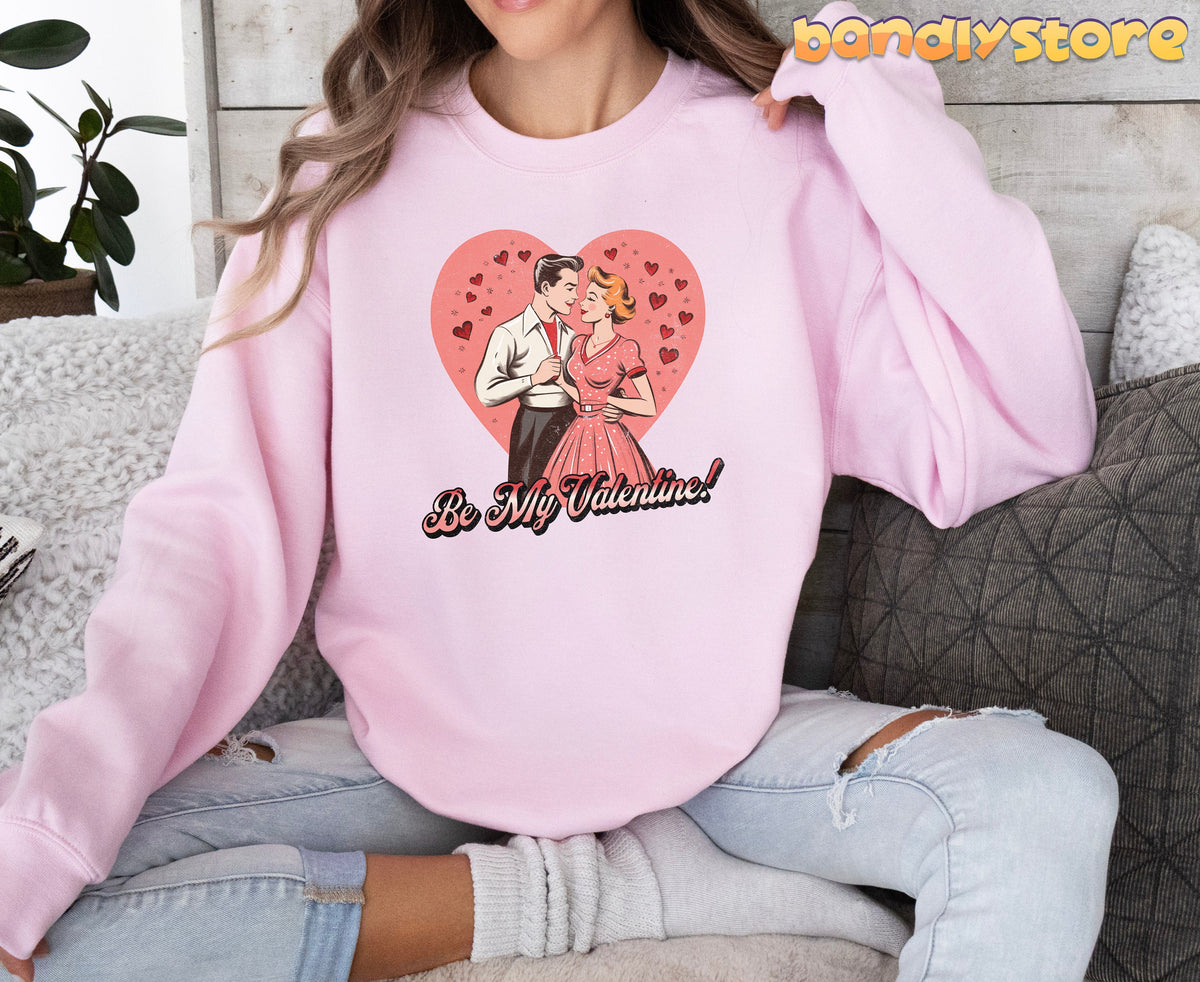 Be Mine Valentine Sweatshirt in light pink worn by a person sitting front-facing amid sofa pillows and a plant.