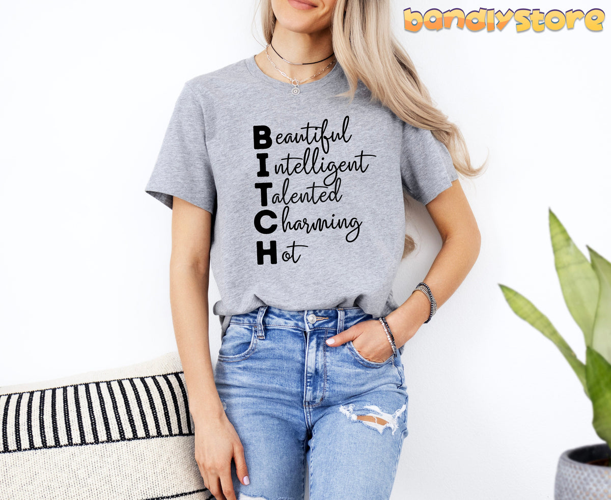 Bitch Definition T-Shirt in Athletic Heather front view on model by white wall with striped pillow and plant.