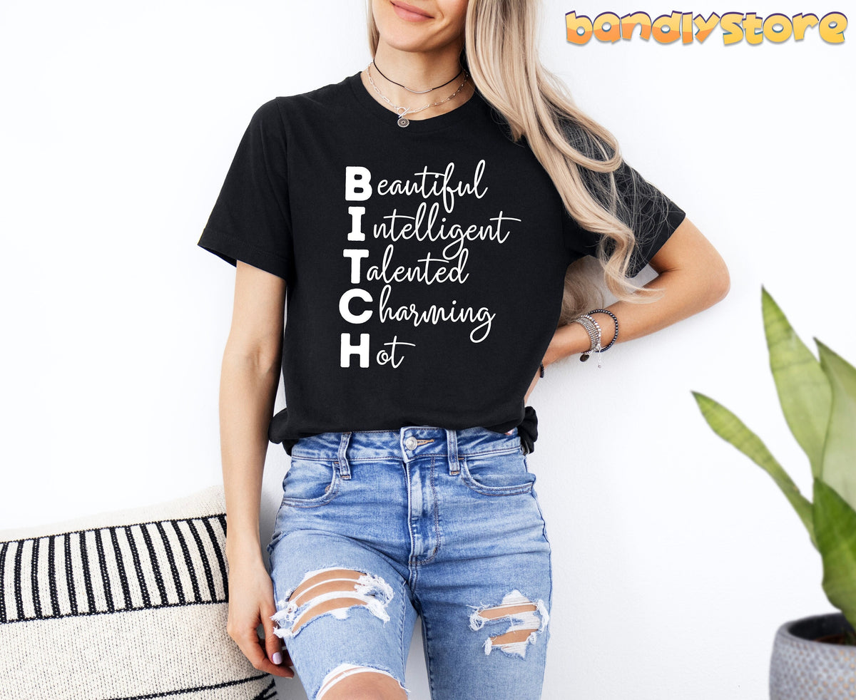 Bitch Definition T-Shirt on a blonde woman in black tee and ripped jeans, leaning by a white wall with pillow and plant.