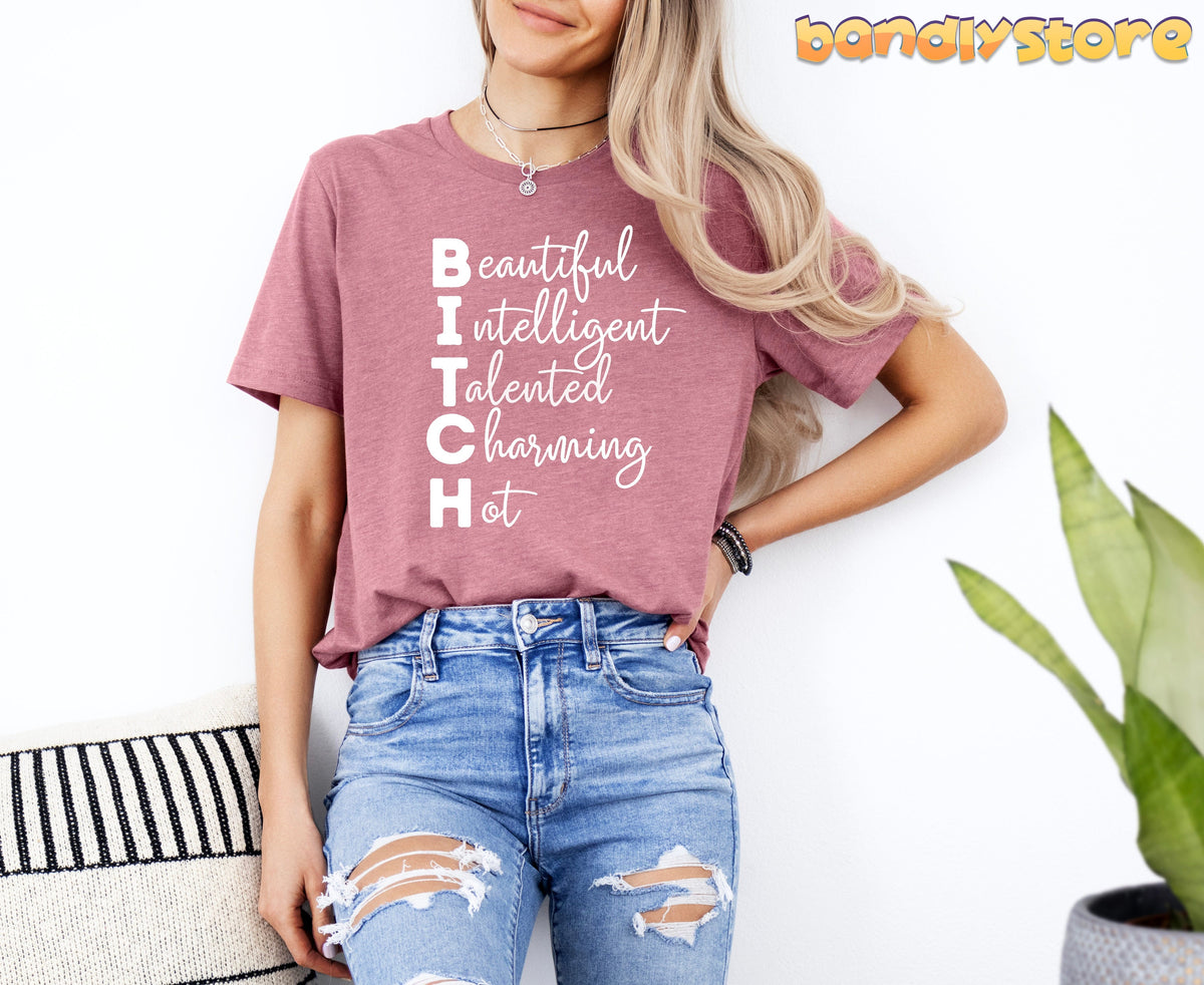 Bitch Definition T-Shirt in Heather Mauve on a woman leaning against a white wall beside a plant and striped pillow.