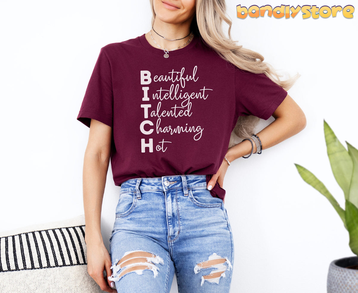 Bitch Definition T-Shirt in maroon on model facing forward against white wall by plant and cushion.