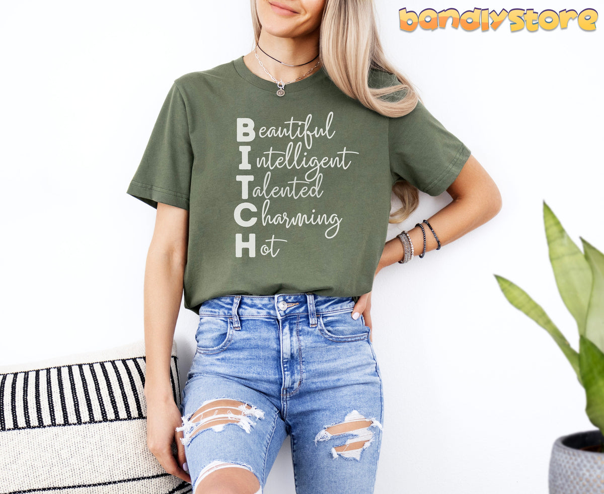 Bitch Definition T-Shirt in Military Green on a model leaning against a white wall beside a striped cushion and plant.
