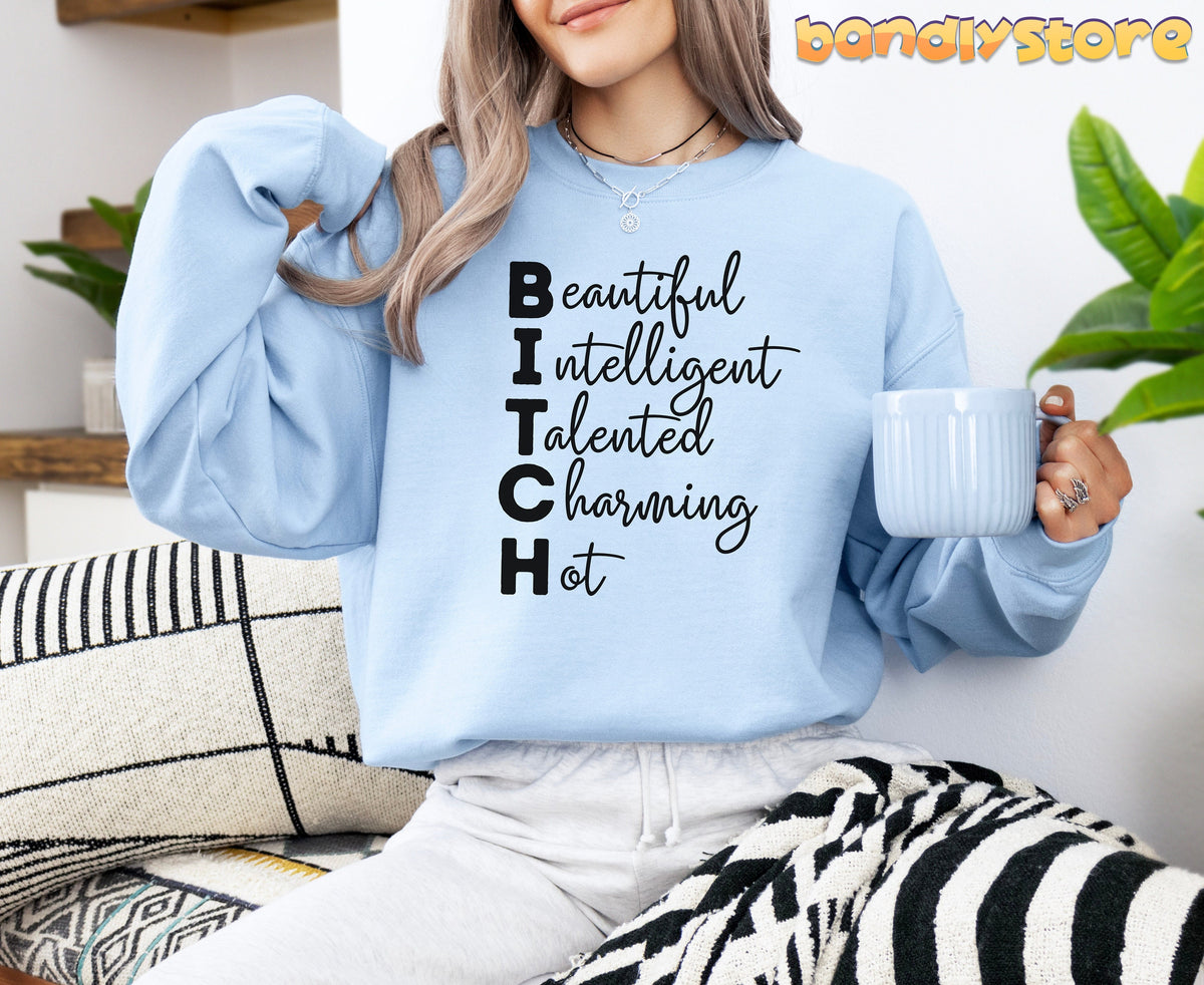 Bitch Definition Sweatshirt in light blue worn by a woman seated on a couch holding a mug amid pillows and plants.