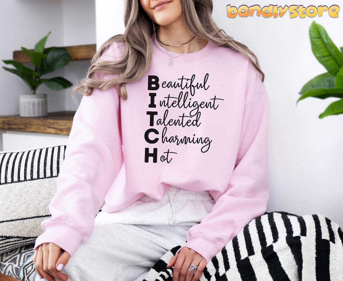 Bitch Definition Sweatshirt in light pink on a seated woman by plants and patterned cushions.