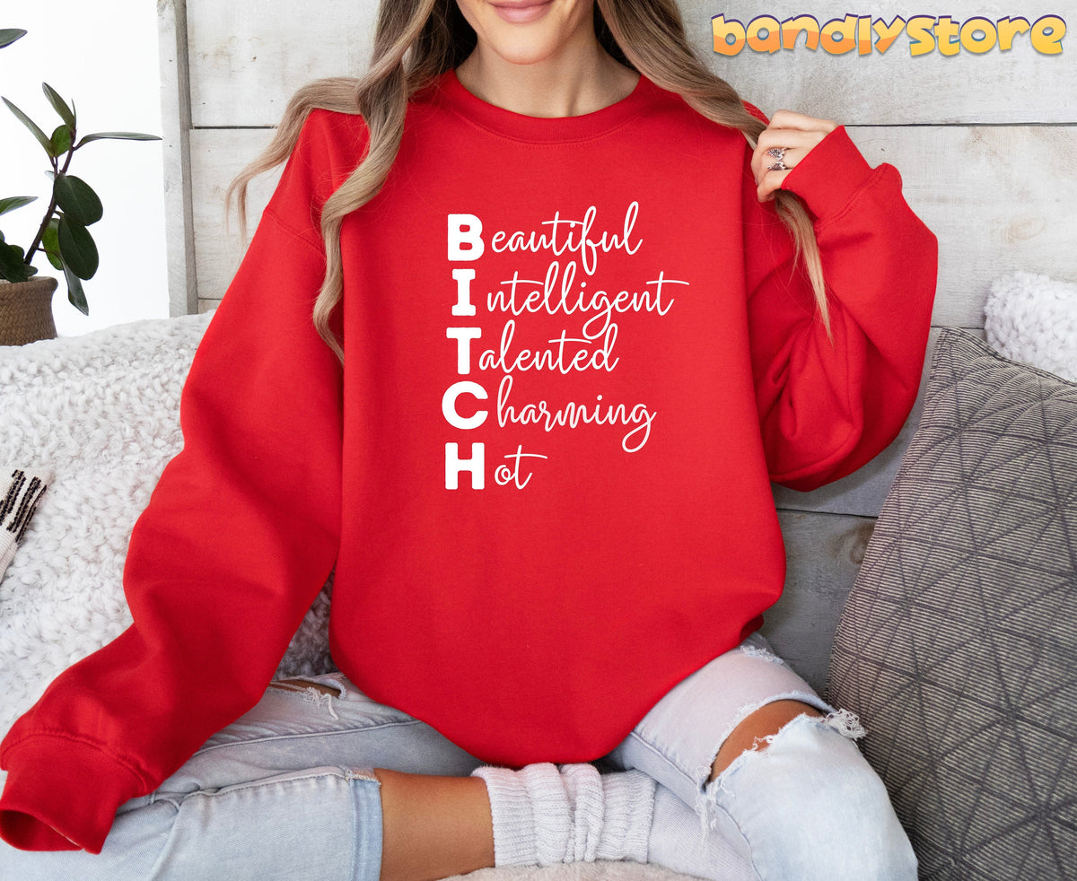 Bitch Definition Sweatshirt in red on seated woman by wood wall with plant and pillows.