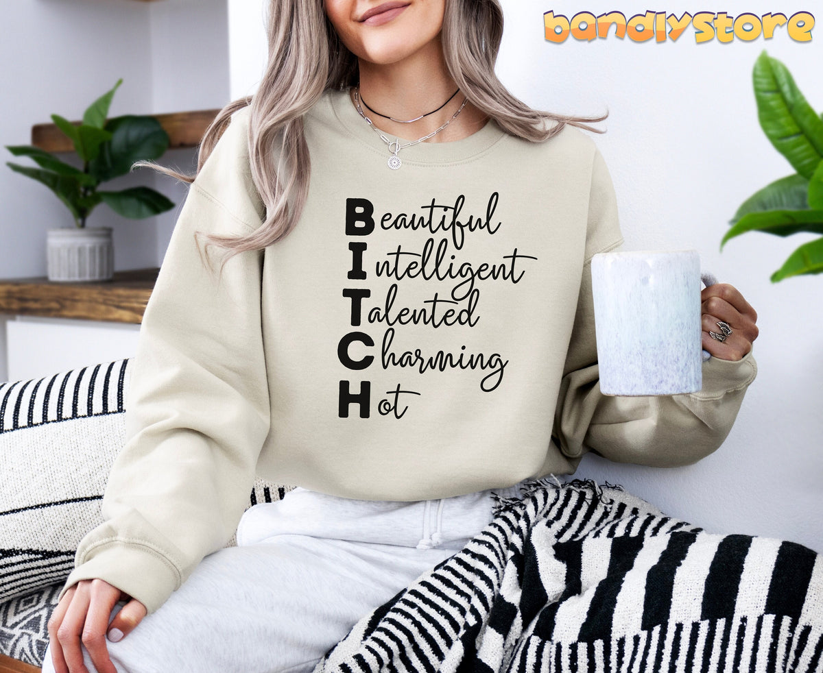 Bitch Definition Sweatshirt in sand, front view on a seated person holding a mug by plants and striped pillows.