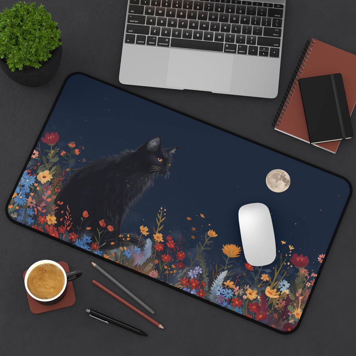 Black Cat And The Moon Desk Mat in 12 × 22 inches, overhead on a desk with laptop, plant, notebooks, coffee and pens.