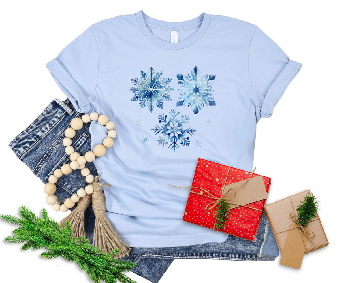 Watercolor Snowflakes T-Shirt in light blue flat lay with jeans, bead garland, greenery and gifts.