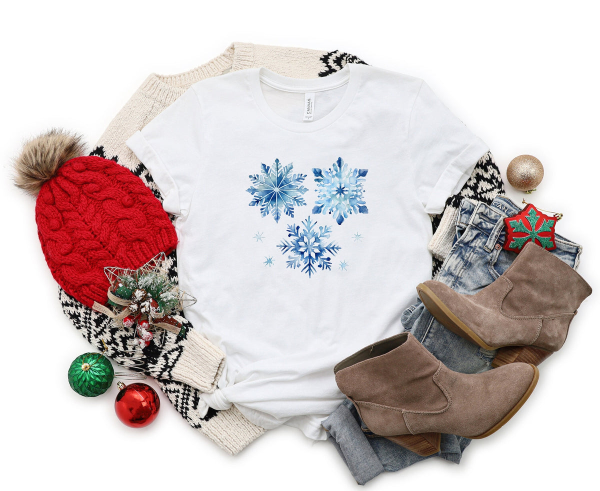 Watercolor Snowflakes T-Shirt in white, flat-laid amid holiday knits, red pom beanie, jeans, boots and ornaments.