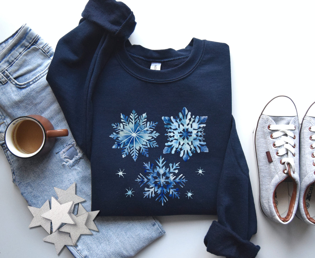 Blue Watercolor Snowflakes Sweatshirt in navy, flat-laid with jeans, coffee mug, wooden stars and sneakers.