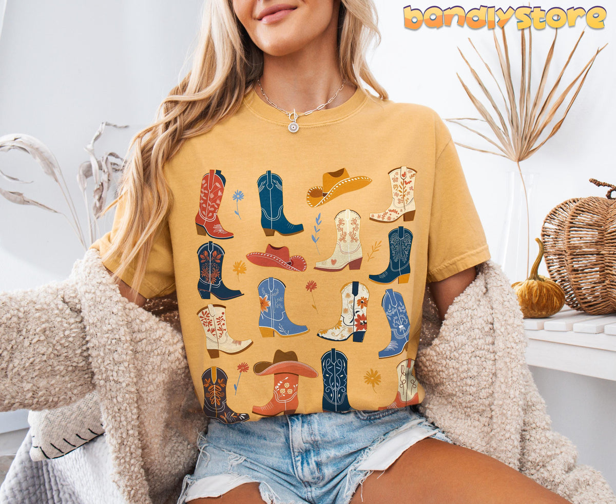 Boho Cowgirl Boots T-Shirt mustard tee worn front-on indoors among dried plants, pumpkins and a woven bag.