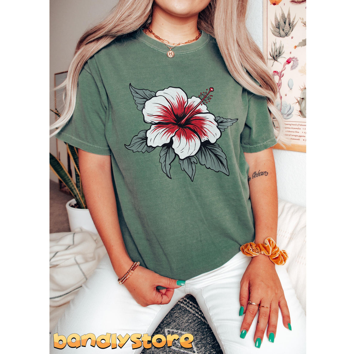 Boho Hibiscus Flower T-Shirt in moss, worn front-facing indoors by a seated person amid plants and a poster.
