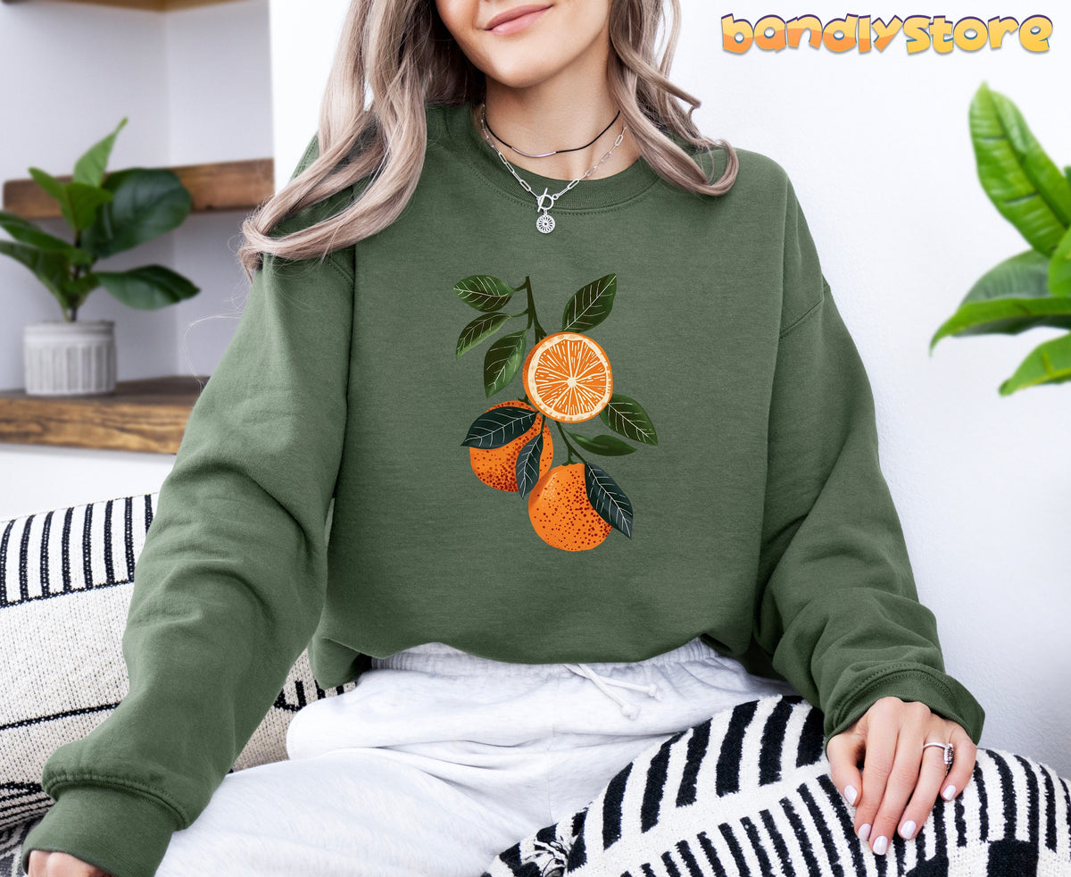 Boho Oranges Sweatshirt in military green, worn seated indoors by shelves, plants, and striped cushions.
