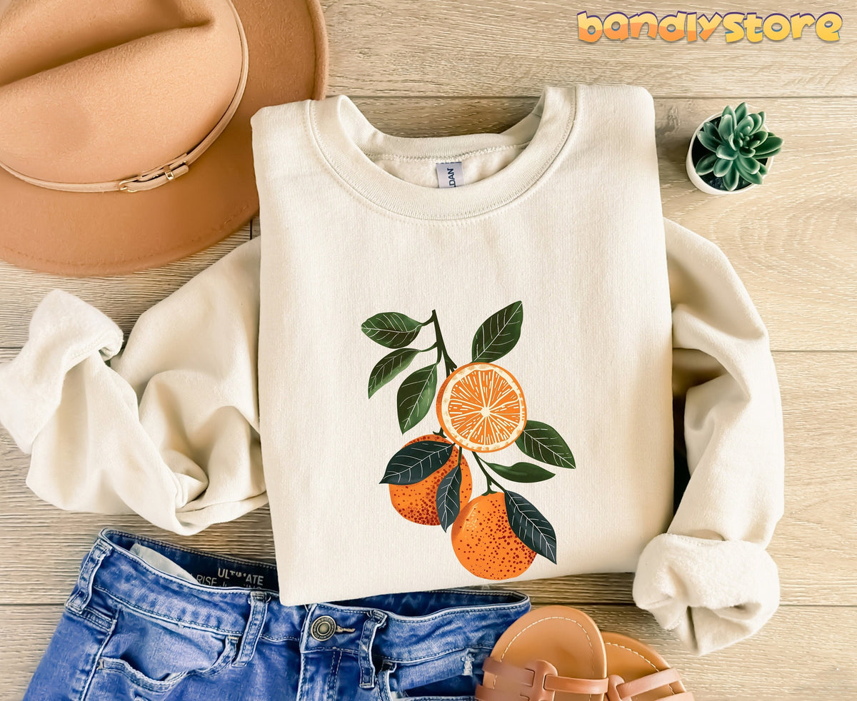 Boho Oranges Sweatshirt in sand flat-laid on wood with hat, jeans, sandals and plant.