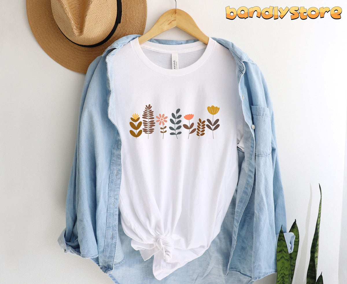 Cute Boho Wildflowers T-Shirt white hanging on a wooden hanger over a denim shirt next to a straw hat and plant.