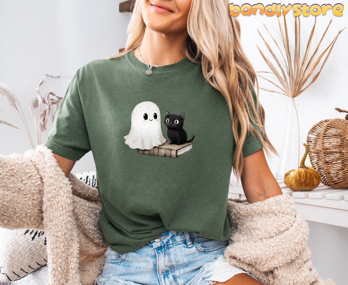 Cute Bookish Ghost T-Shirt in moss shown front-on on a seated model amid dried plants, a pumpkin and woven basket.