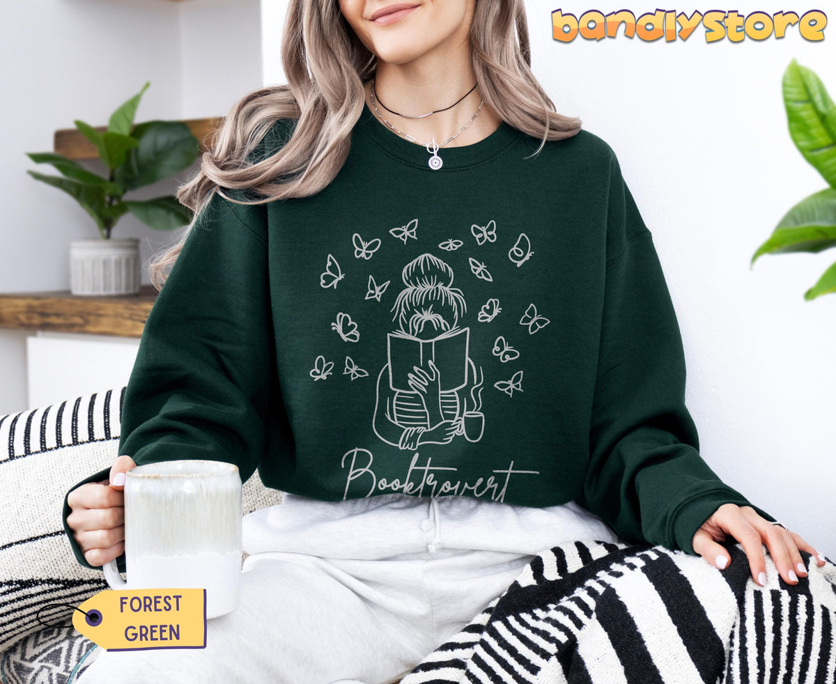 Booktrovert Sweatshirt in forest green, worn seated indoors holding a mug, with plants and striped blanket nearby.