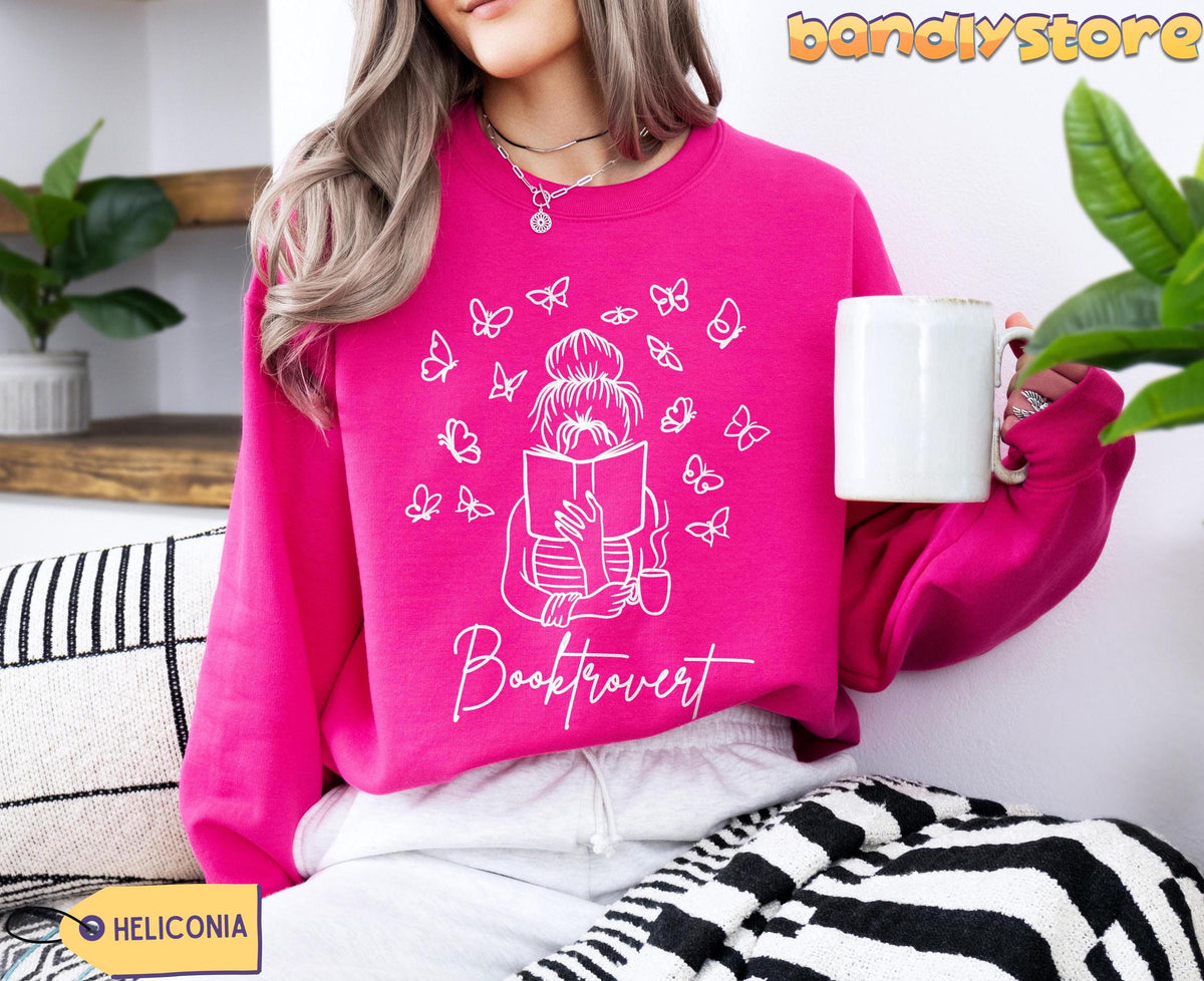 Booktrovert Sweatshirt in Heliconia on a seated person holding a mug near plants and striped pillows.