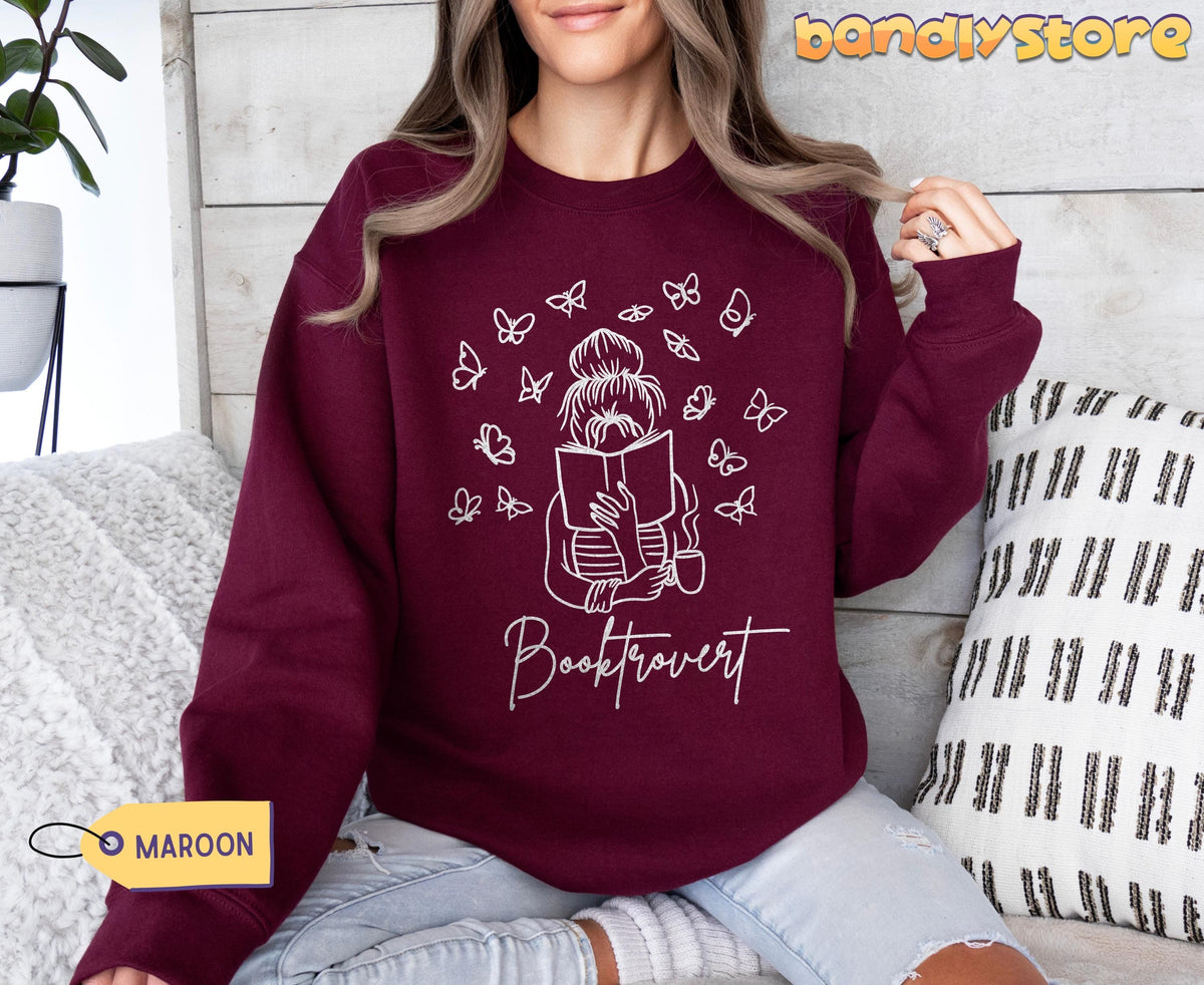 Booktrovert Sweatshirt in maroon on a woman seated facing camera amid pillows and a plant by wooden wall.
