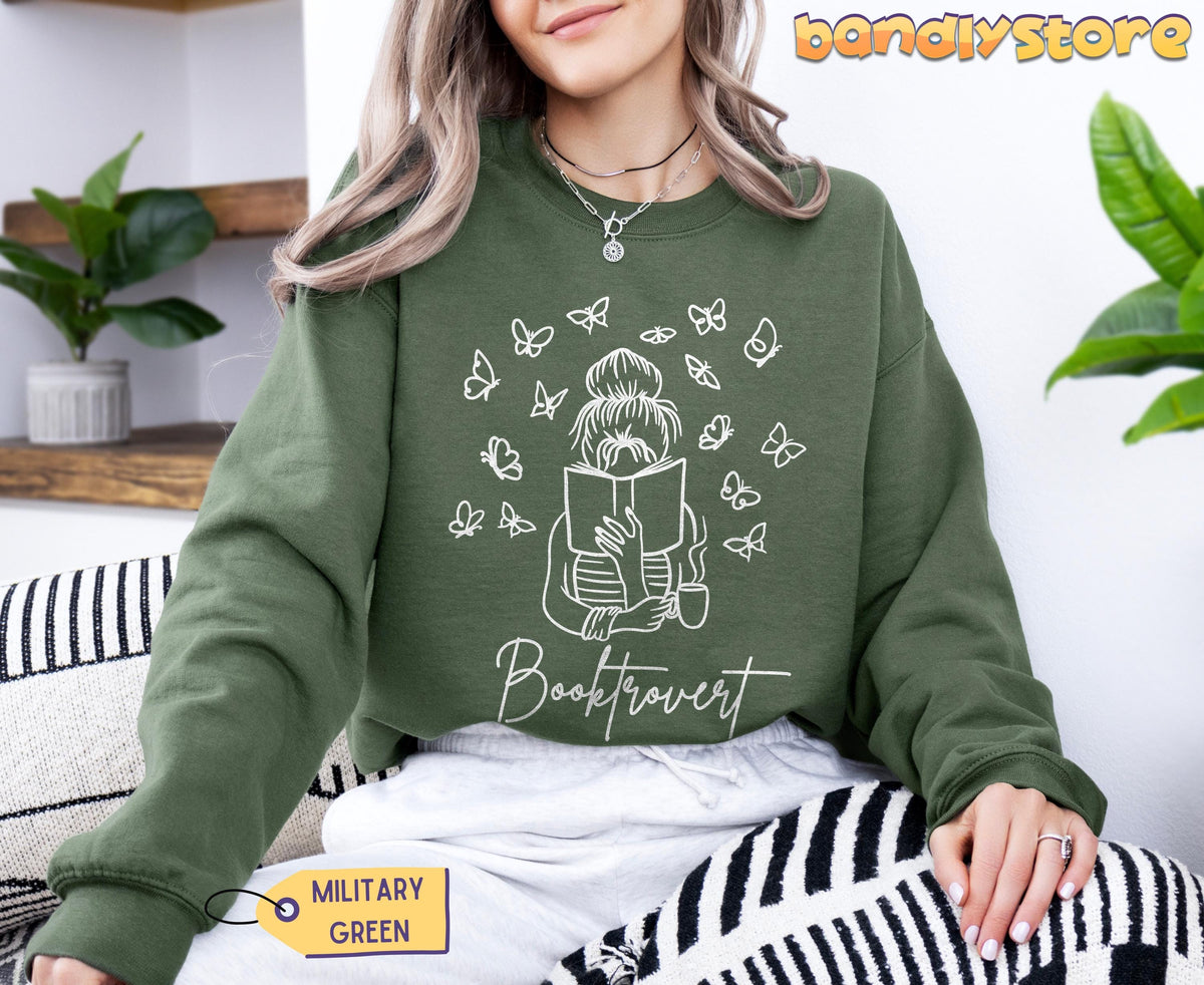 Booktrovert Sweatshirt in military green, front view on a seated person beside plants and patterned cushions.