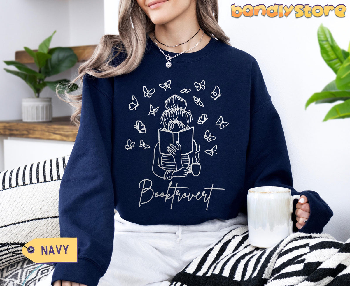 Booktrovert Sweatshirt in navy, worn seated indoors with mug, plants, and striped blanket visible.